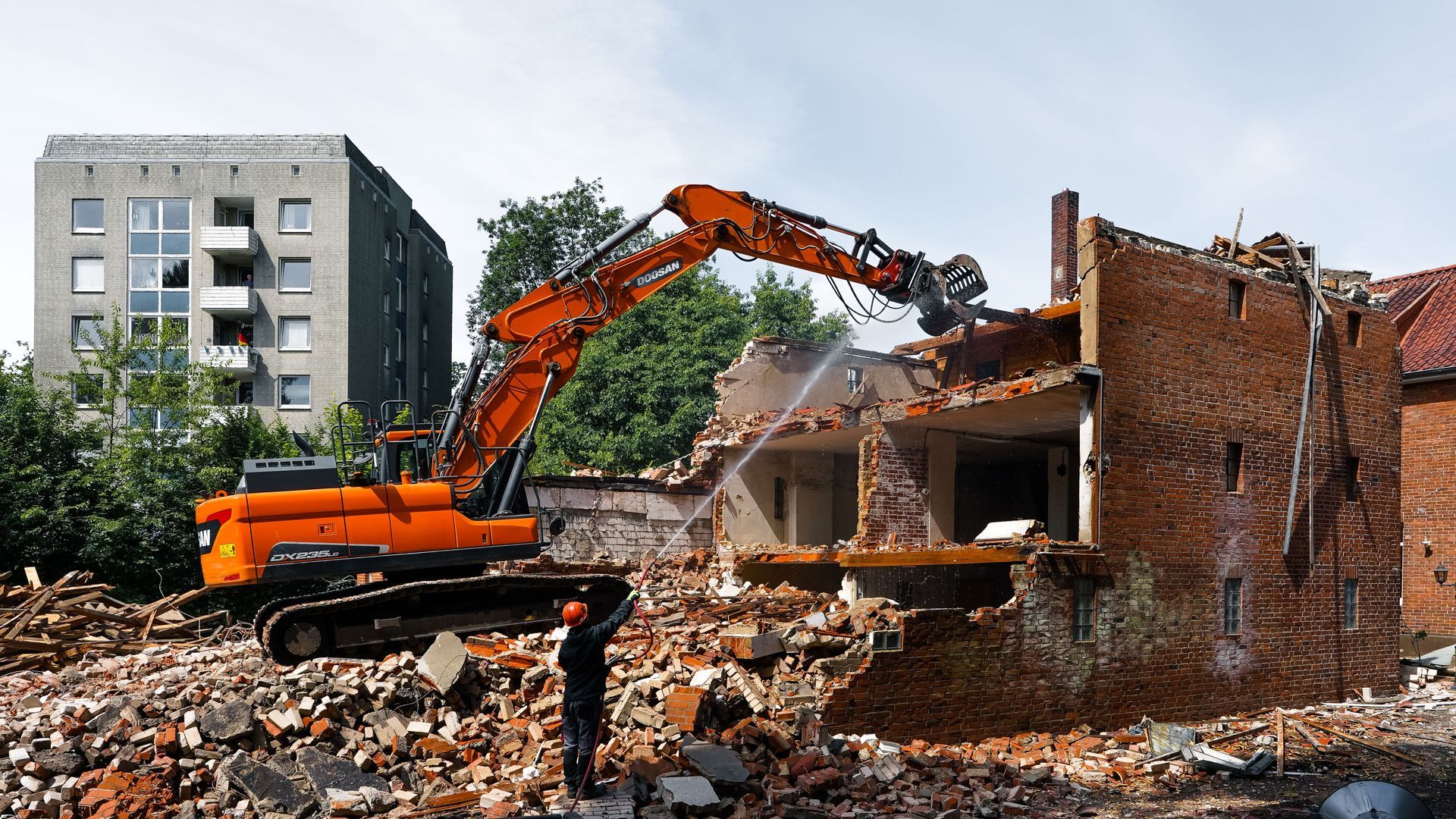 Orange excavator demolishing a brick house amid rubble, with a tall apartment building in the background