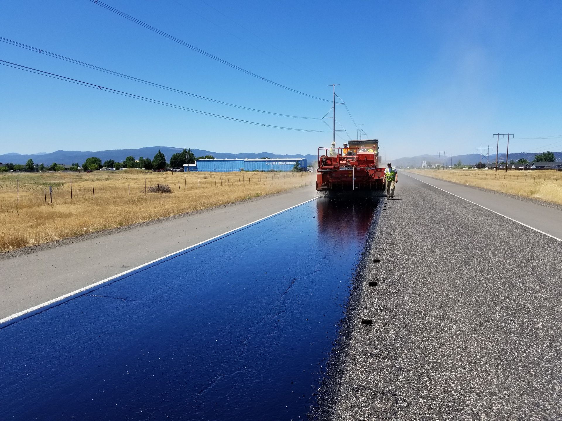 Road crew spraying dark asphalt sealant on a rural highway under a clear blue sky