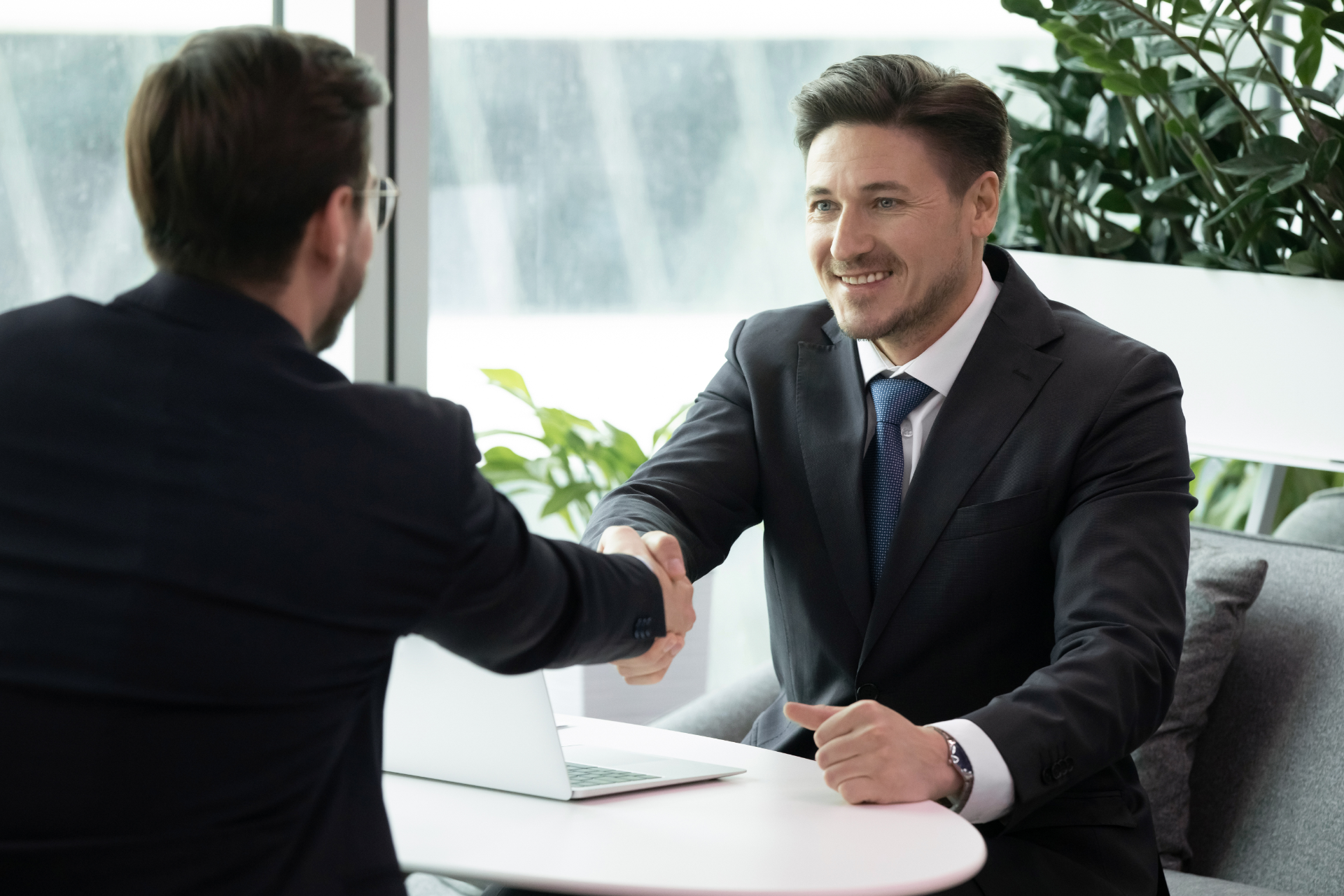 Two businesspeople shake hands across a table in a bright office meeting room.