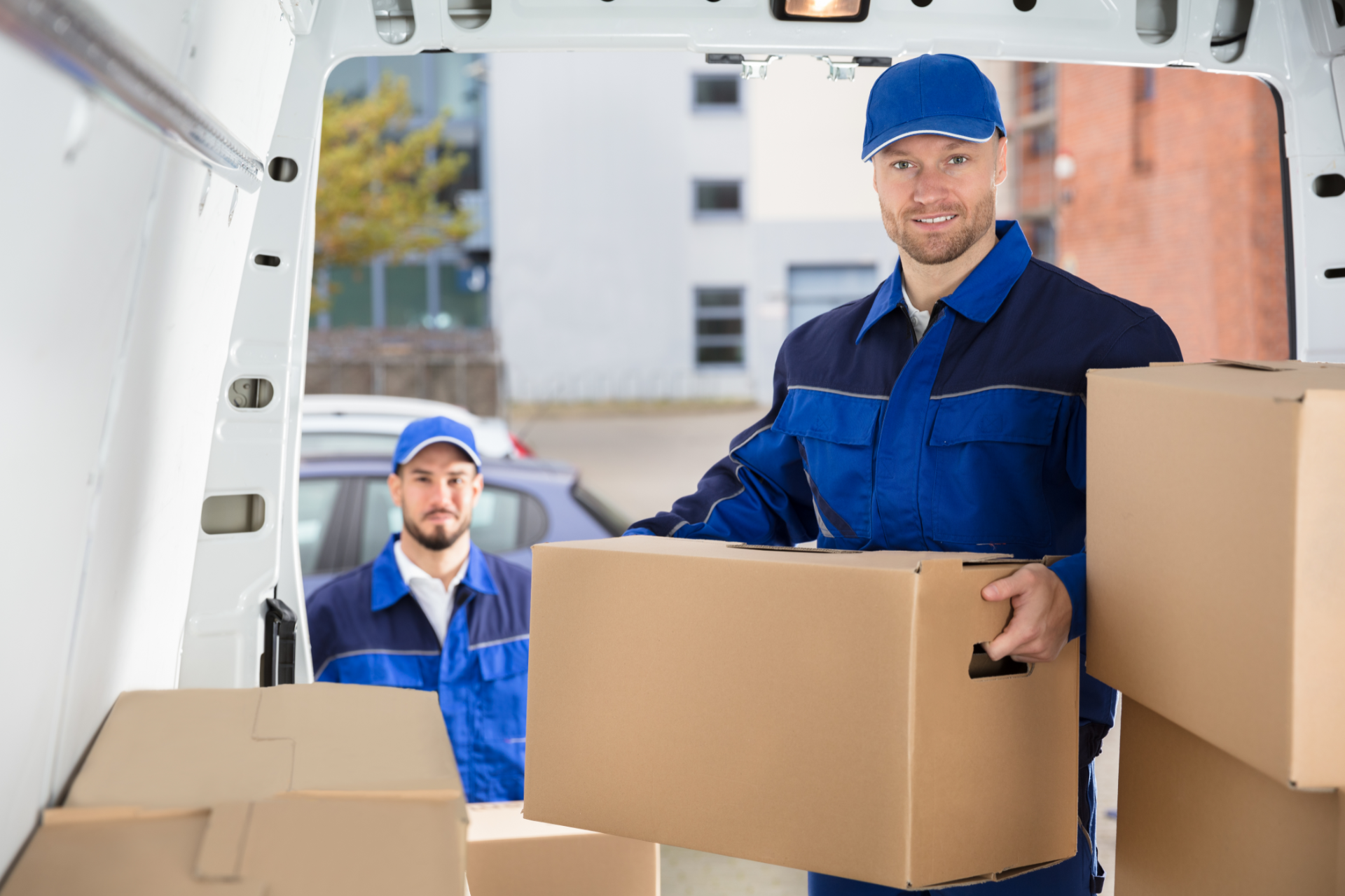 Two movers in blue uniforms carrying boxes from a delivery van near apartment buildings