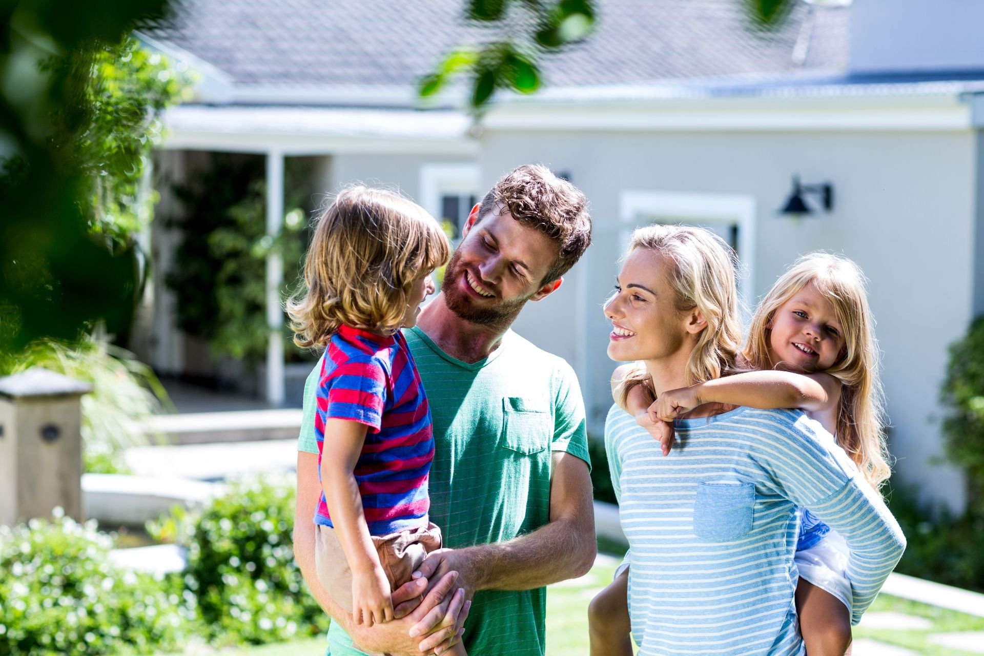 Family smiling in a sunny yard, with a child on each parent's back in front of a house.
