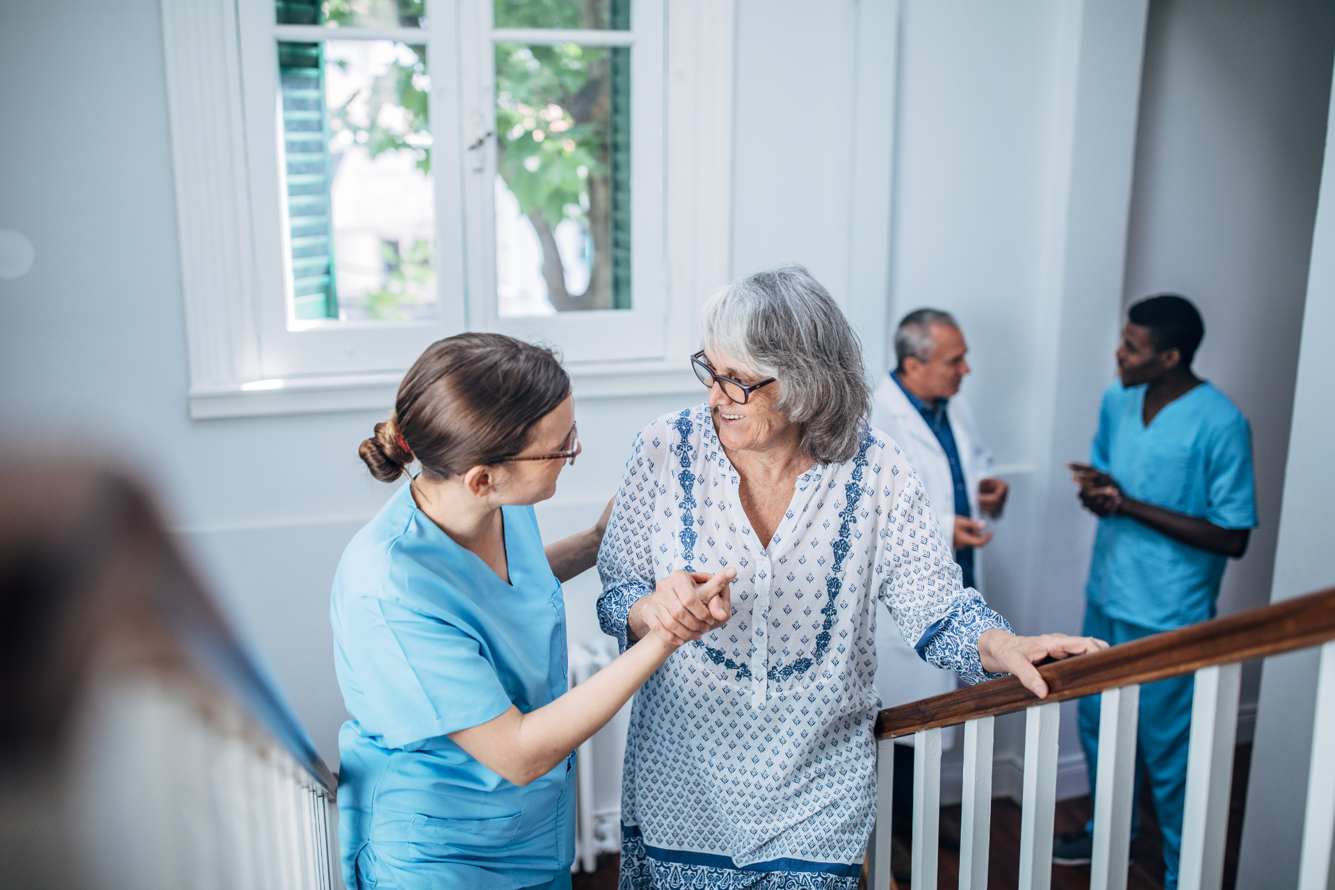 Nurse assisting a patient on a stairway in a bright care facility, with two staff members in the background