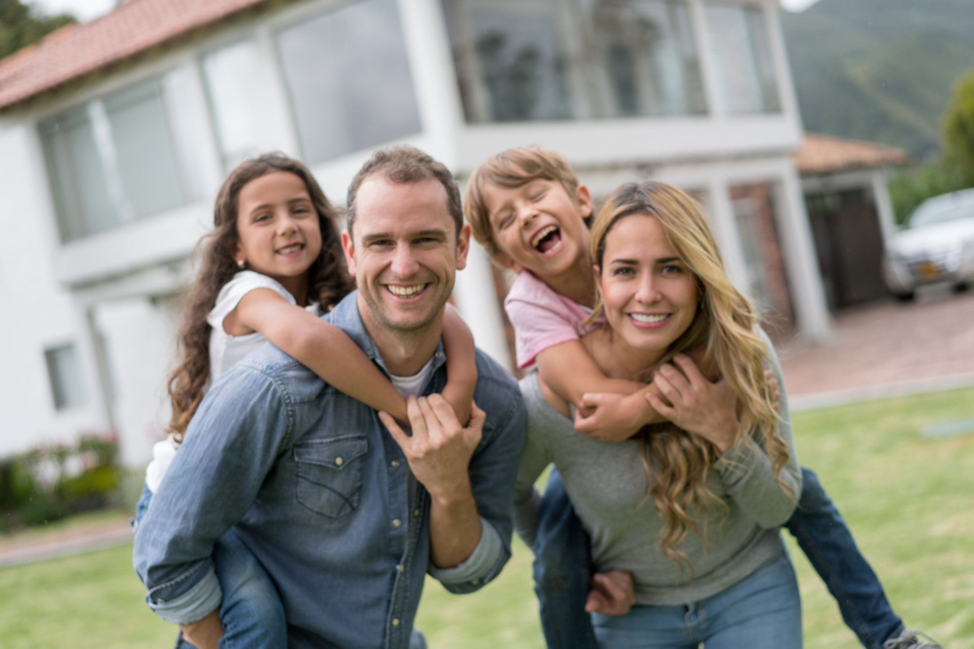 Smiling family of four posing outdoors in front of a modern house, two children on parents’ backs.