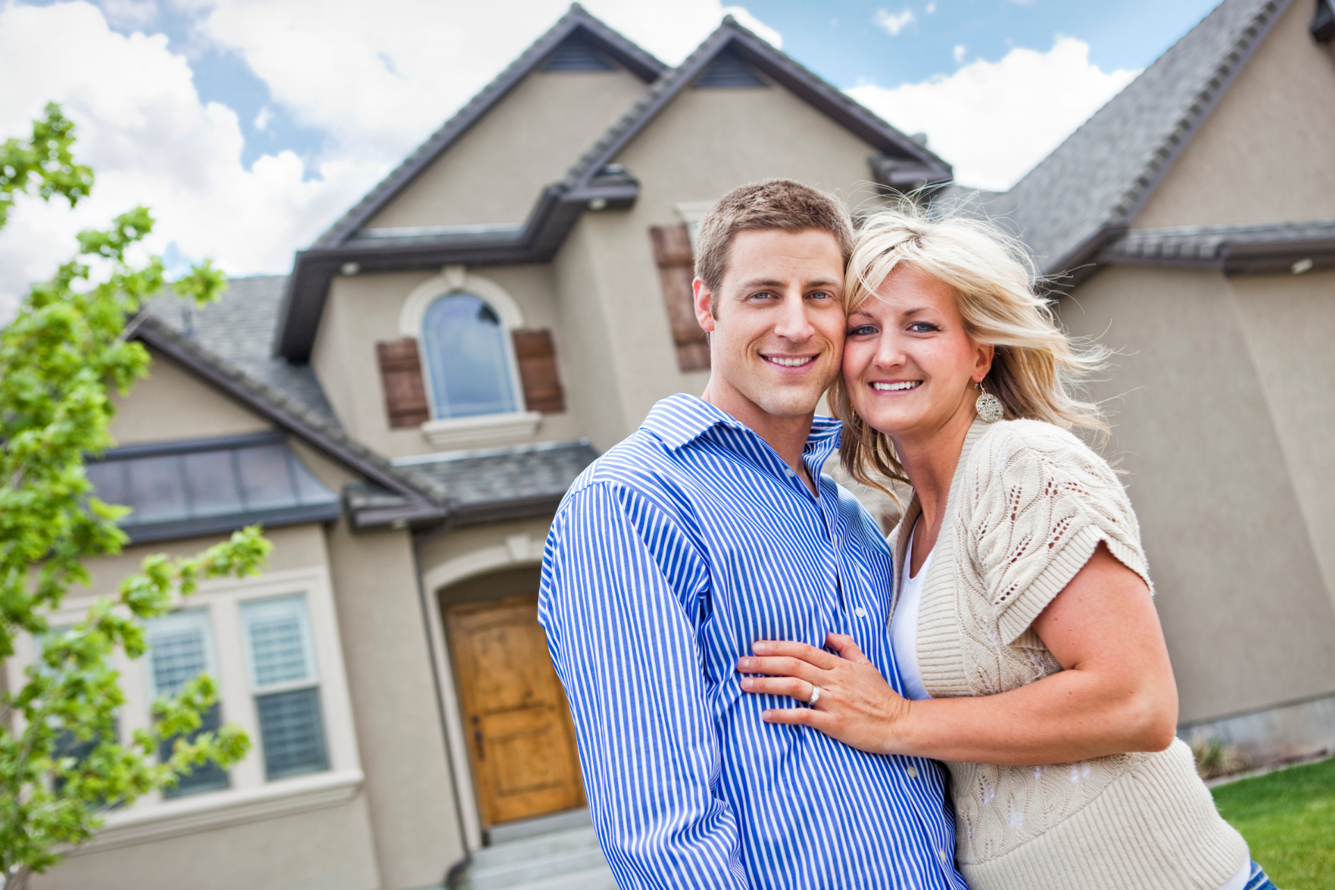 Smiling couple standing in front of a house, with the man in a blue striped shirt and the woman in a beige top.