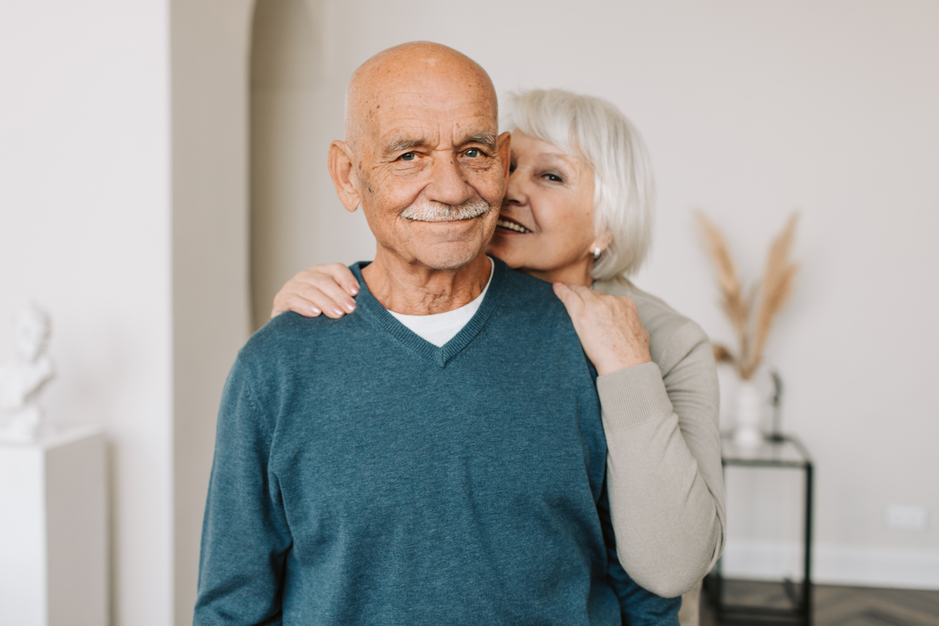 Smiling couple embracing in a bright home interior, with an elderly man in a teal sweater in front.