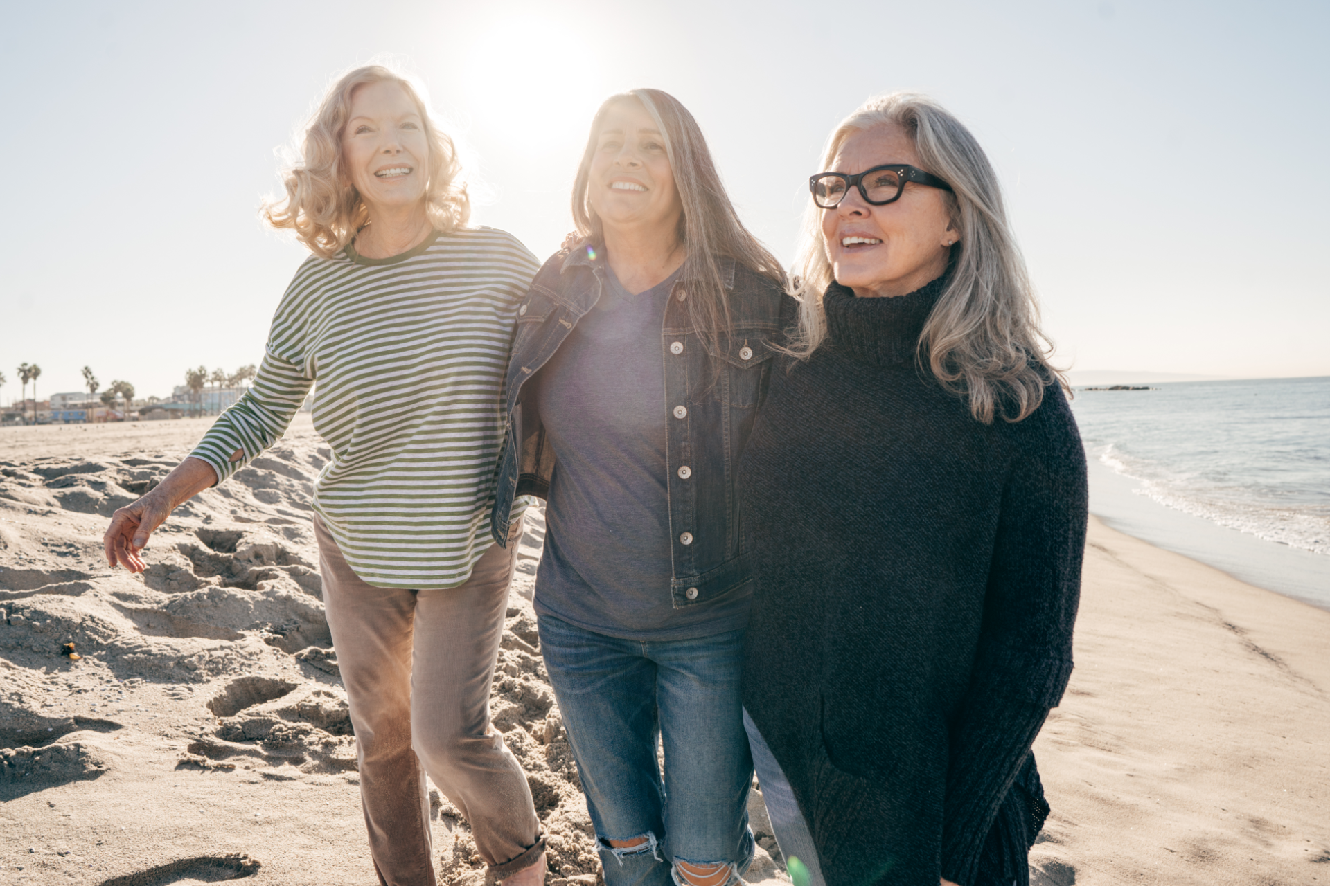Three people walking on a sunny beach, smiling and holding each other close.