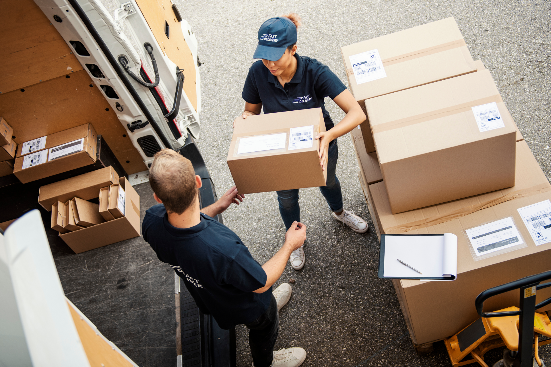 Two workers in a storage room hand off a cardboard box among stacked packages.