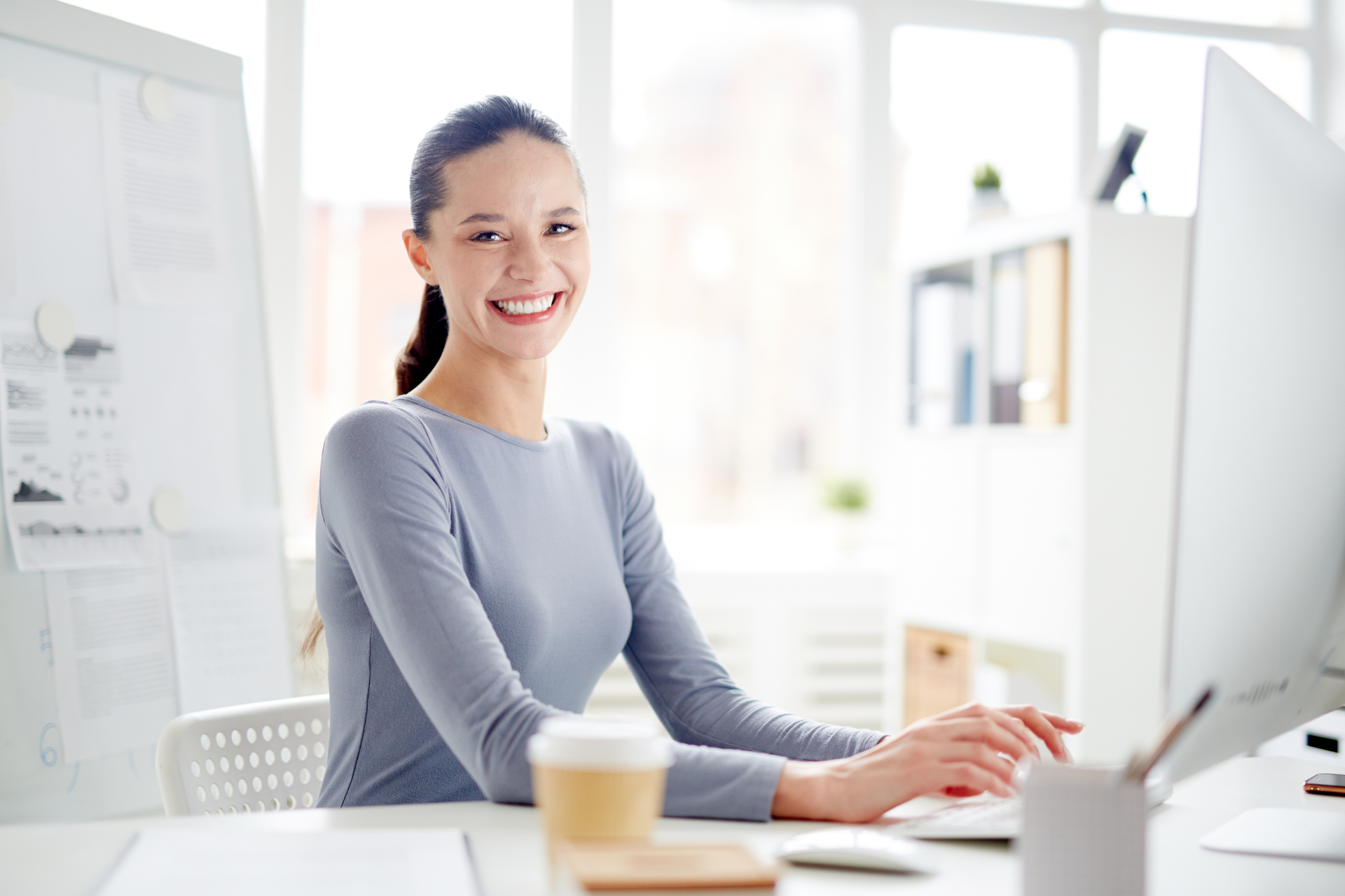 Smiling woman typing at a desk in a bright office, with a coffee cup nearby