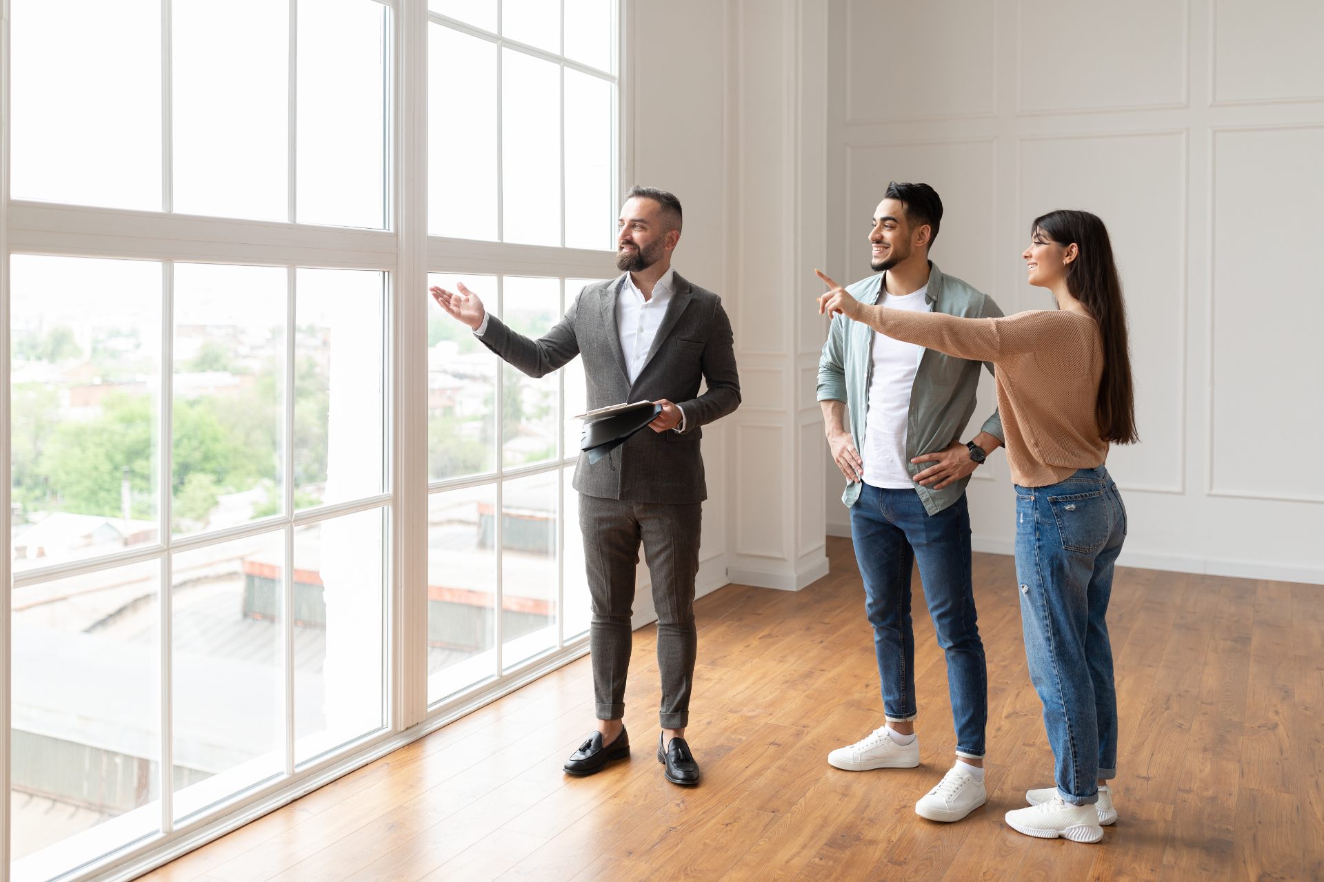 Three people touring a bright empty room, with one man pointing toward large windows.