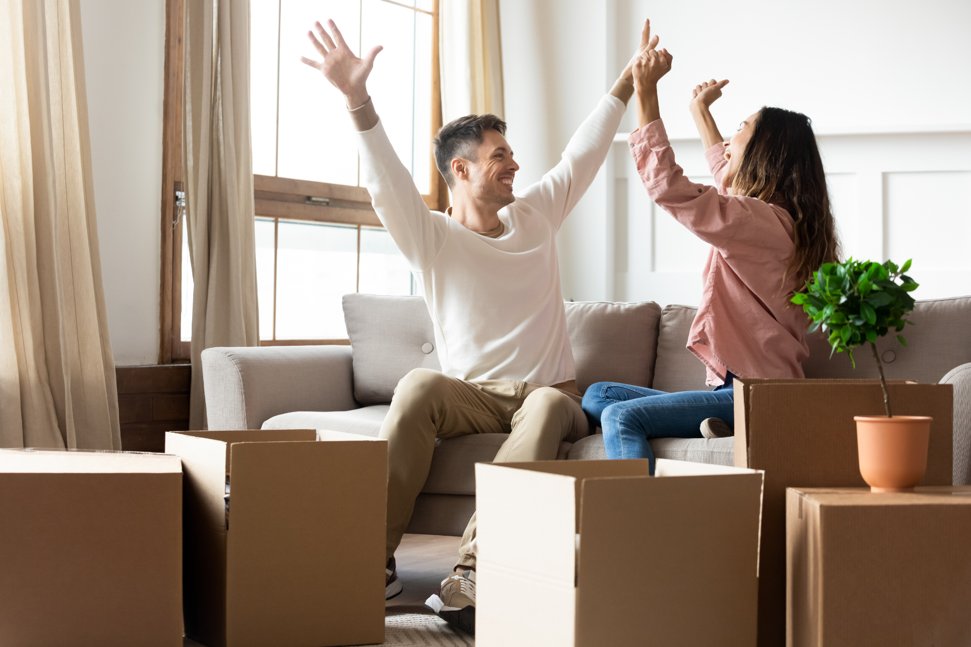Two people celebrating on a couch amid moving boxes in a bright living room