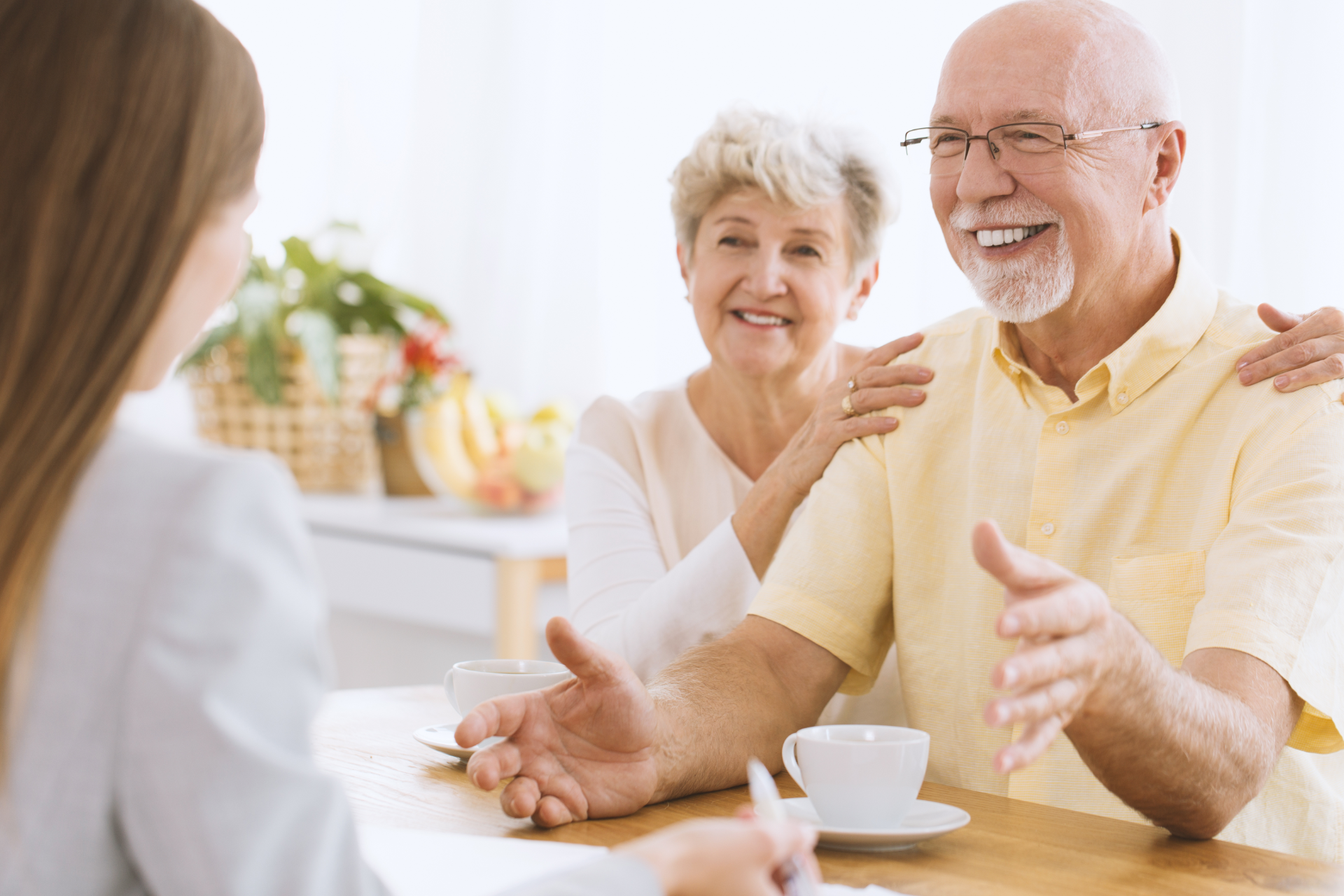 Two smiling people chatting at a café table, with cups and a bright window in the background