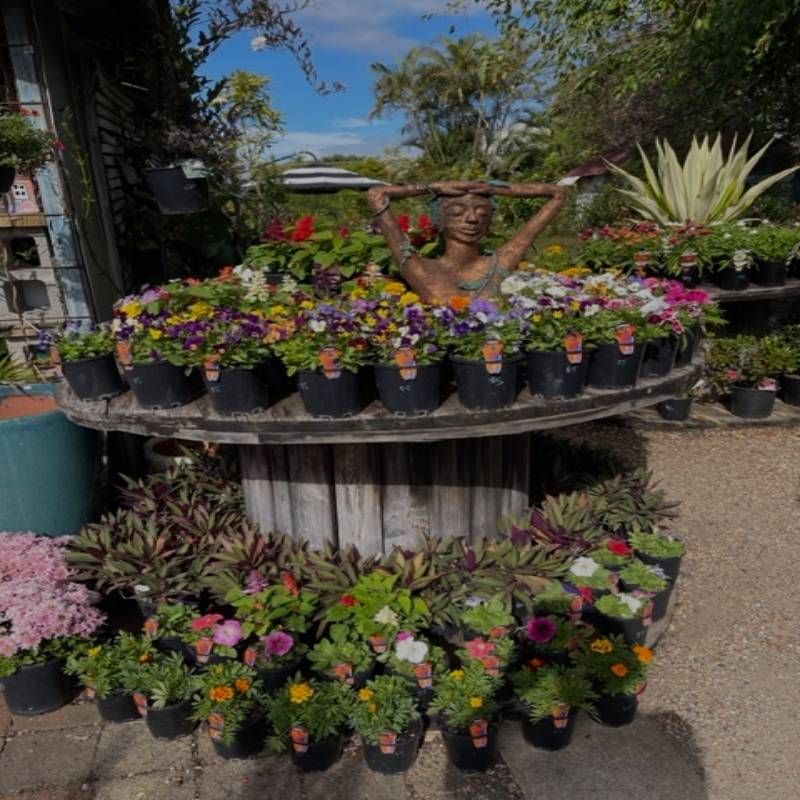 A wooden cable reel overflowing with potted flowers, topped by a bronze statue raising arms, surrounded by plants.