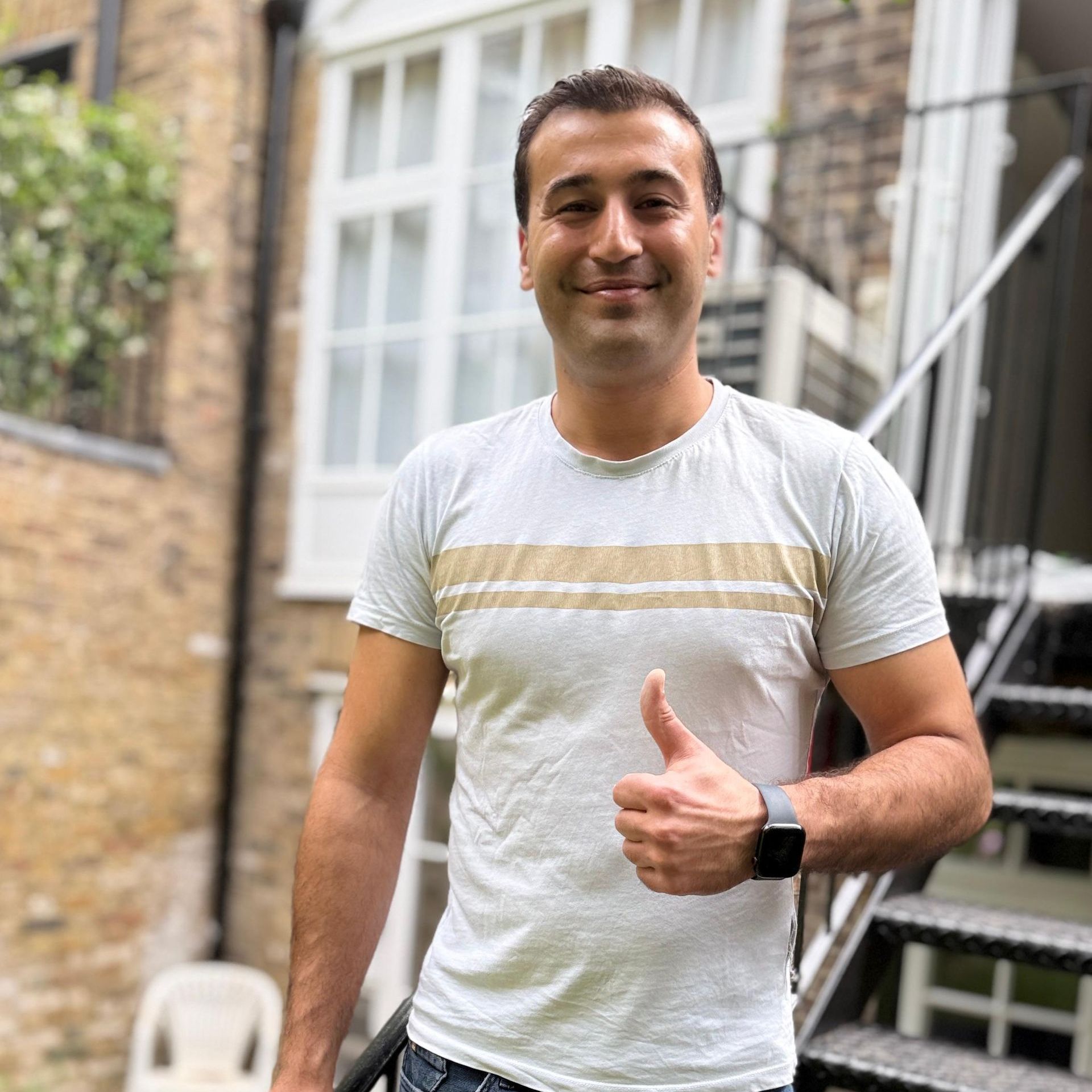 Smiling man in a light striped T-shirt giving a thumbs-up outdoors in a courtyard