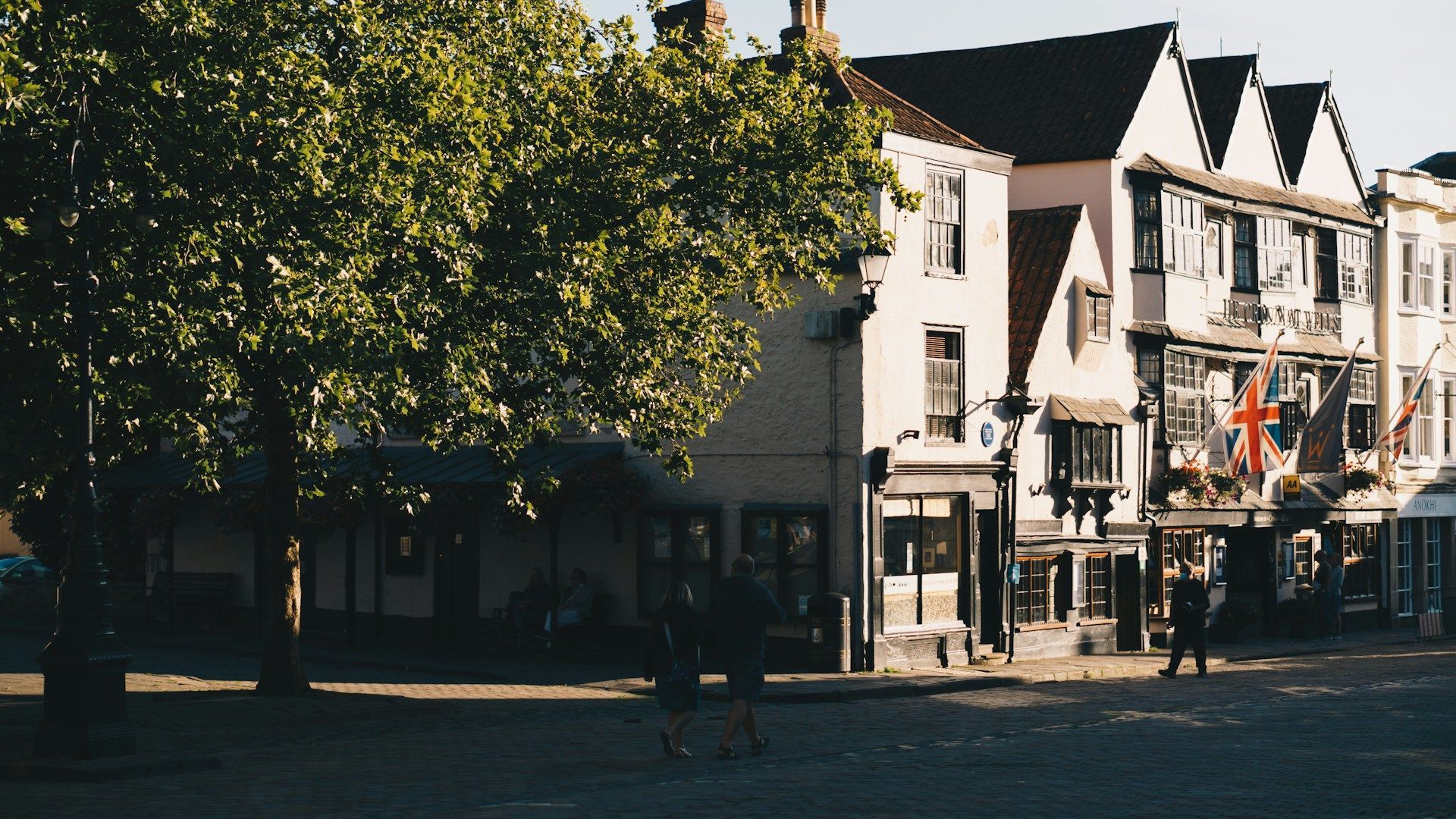 Sunlit town square with trees, historic buildings, and a street café under blue sky