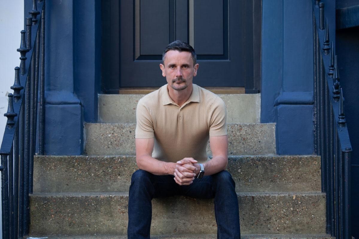 Man sitting on stone steps before a dark blue door, hands clasped, wearing a tan shirt and dark pants