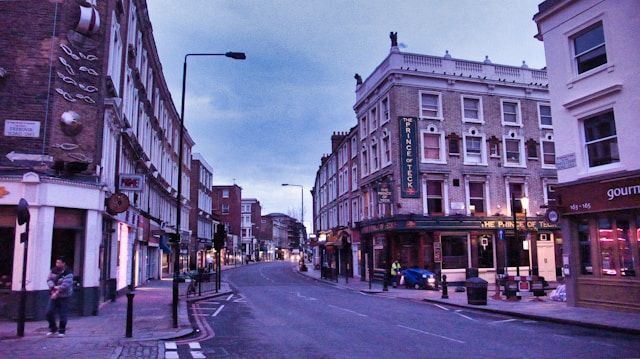 Quiet city street at dusk with historic buildings, shops, and a few pedestrians under a blue sky