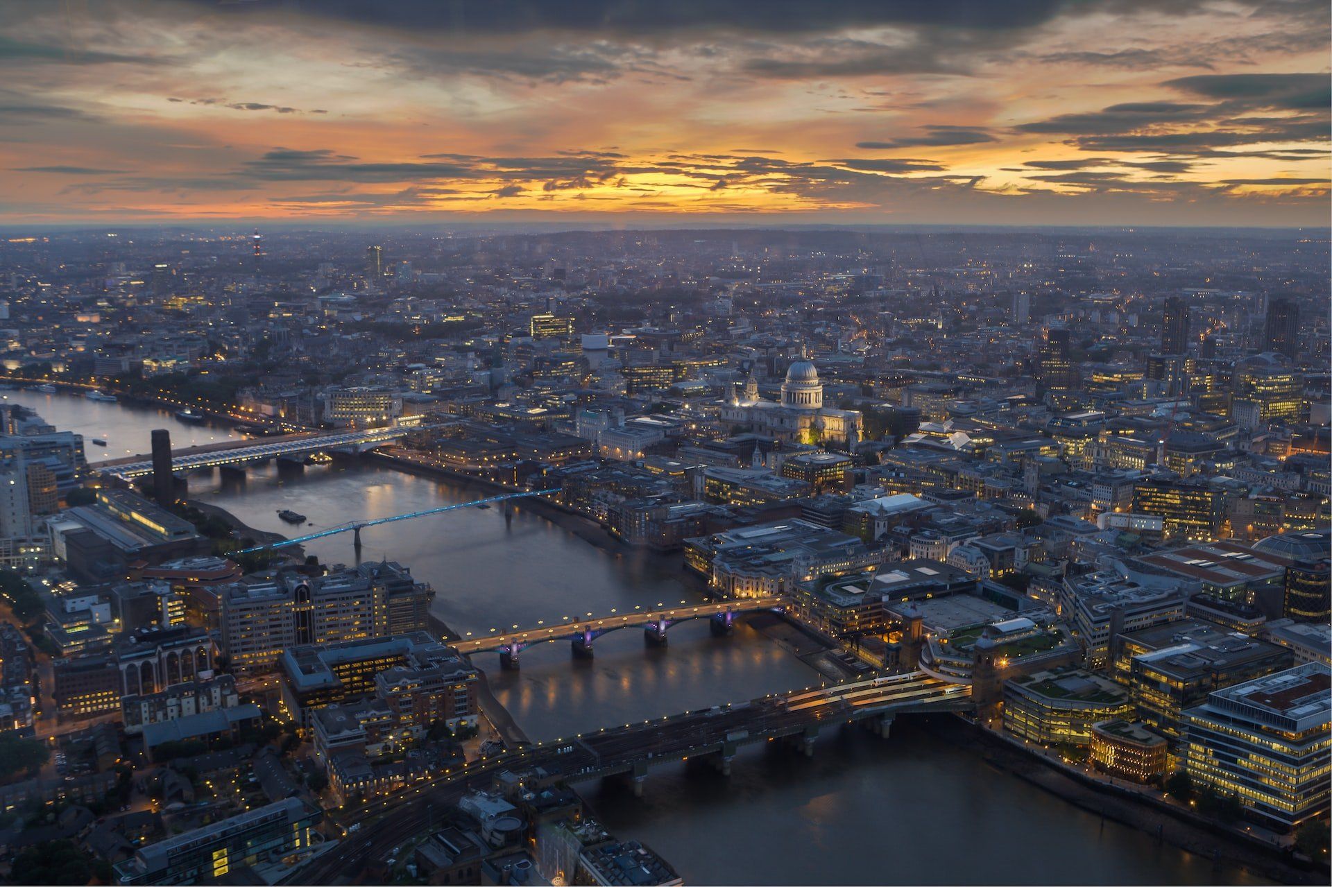 Aerial view of a city at dusk with a river, bridges, and glowing lights under a cloudy sunset sky