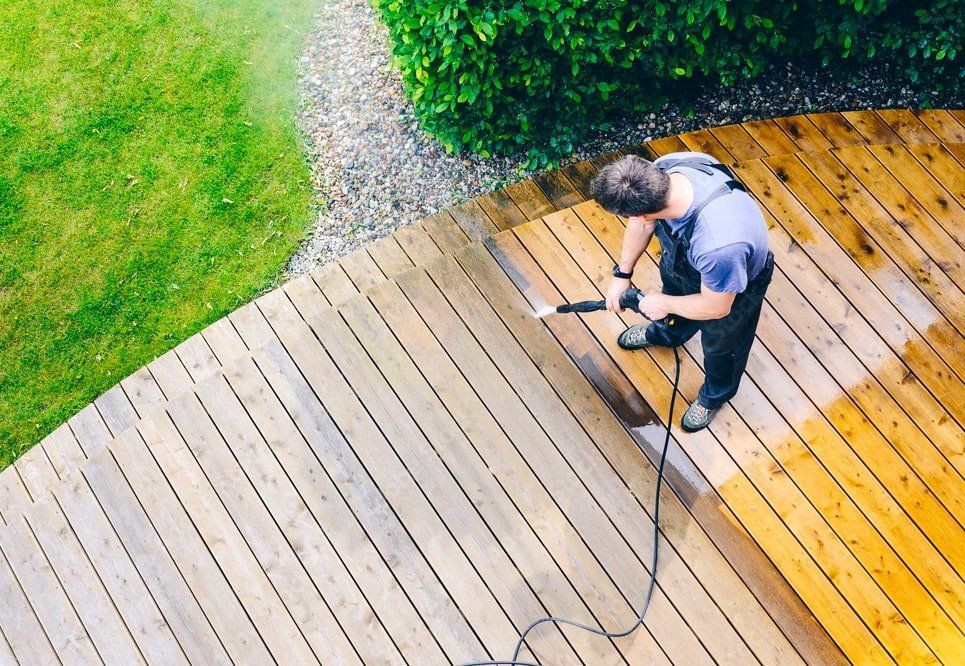 Person pressure washing a wooden deck beside a green lawn and gravel path