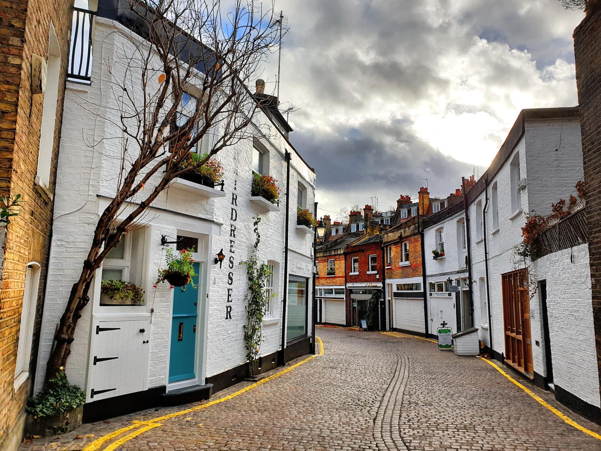 Narrow cobblestone street with white houses, blue door, and cloudy sky in a quaint village.