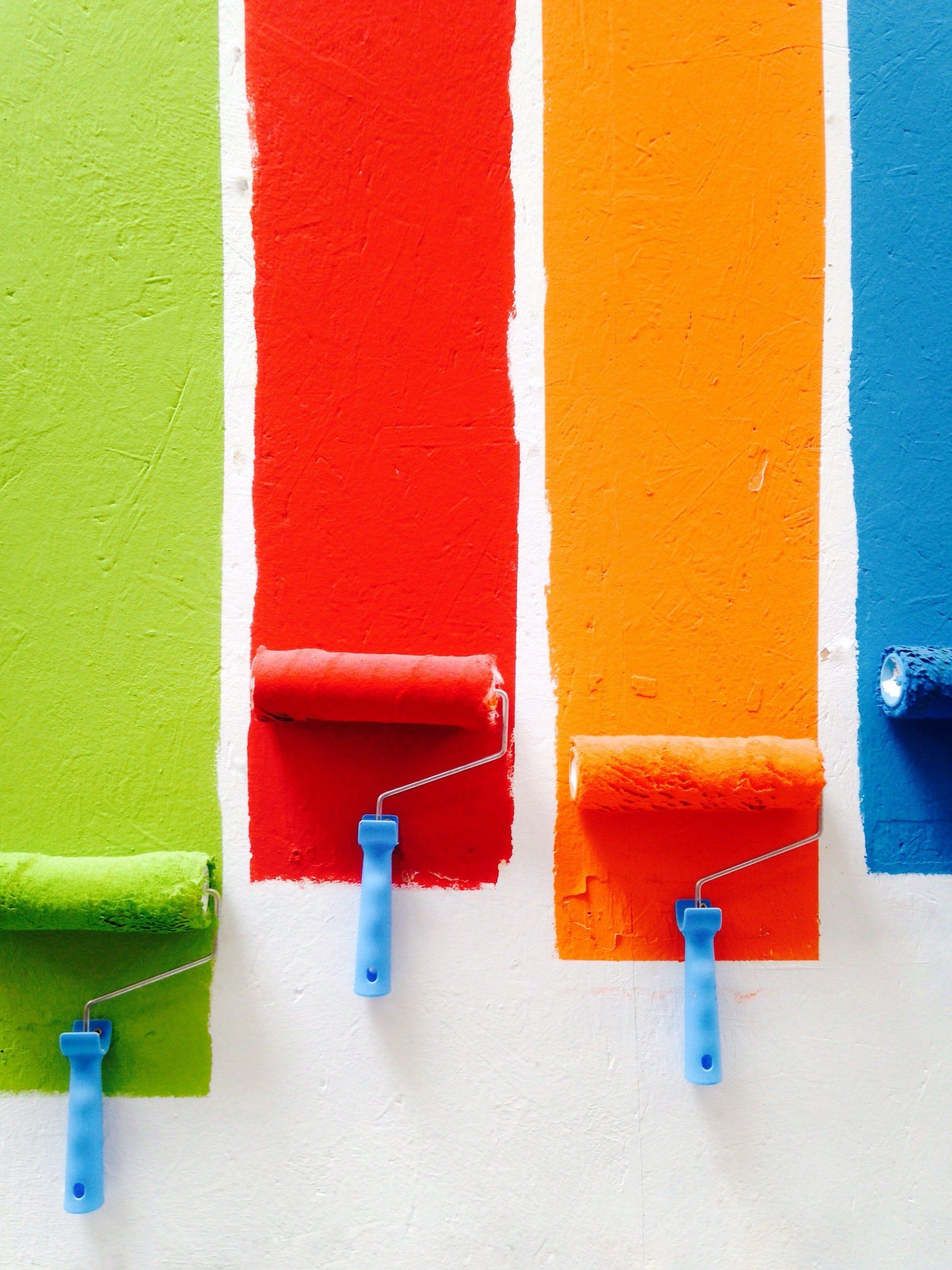 Four colorful paint rollers with drips on striped green, red, orange, and blue paint walls
