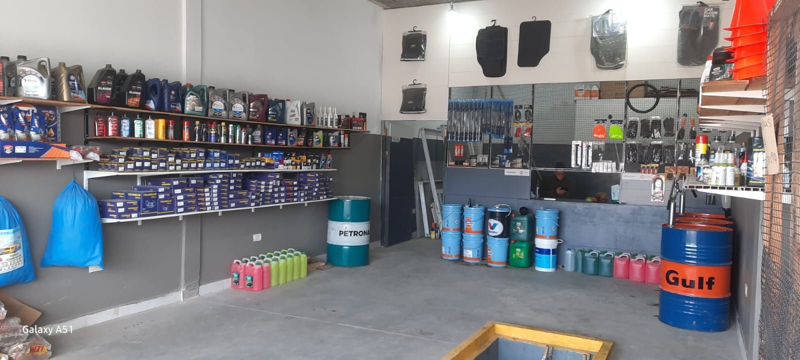 Taller en el garaje con estantes llenos de pintura, herramientas y suministros, además de cubos y un bidón de petróleo Gulf.