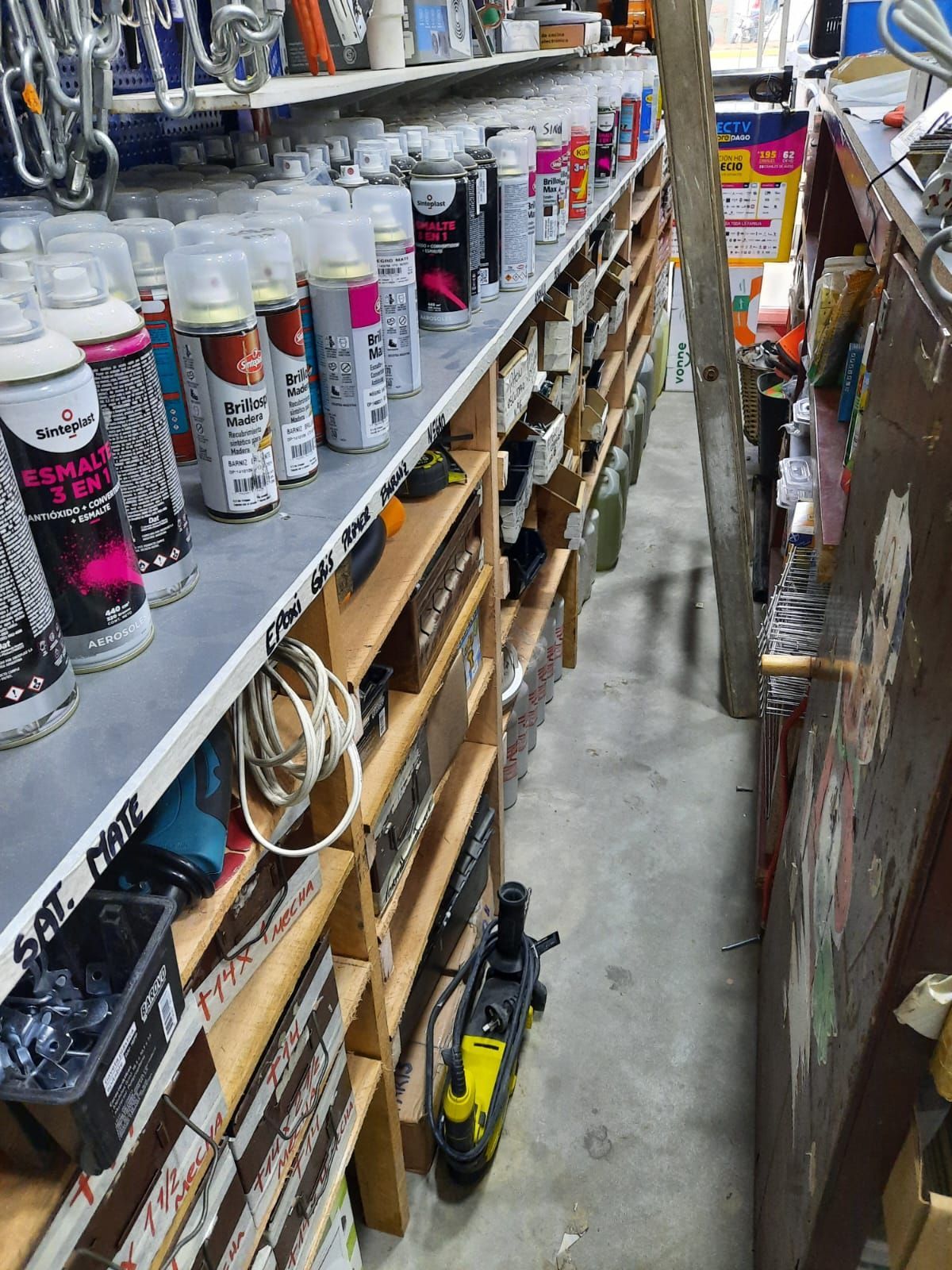 Pasillo estrecho de una ferretería con estantes llenos de latas de aerosol, cajas y herramientas.
