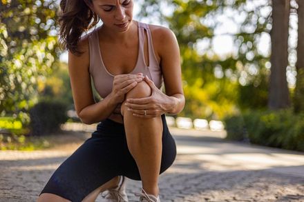 Woman kneeling outdoors and holding her knee, appearing to have a leg injury on a sunlit path