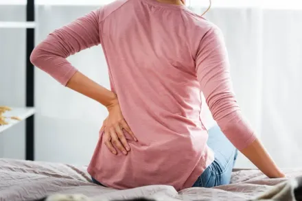 Person sitting on a bed, holding lower back in pain, wearing a pink shirt.