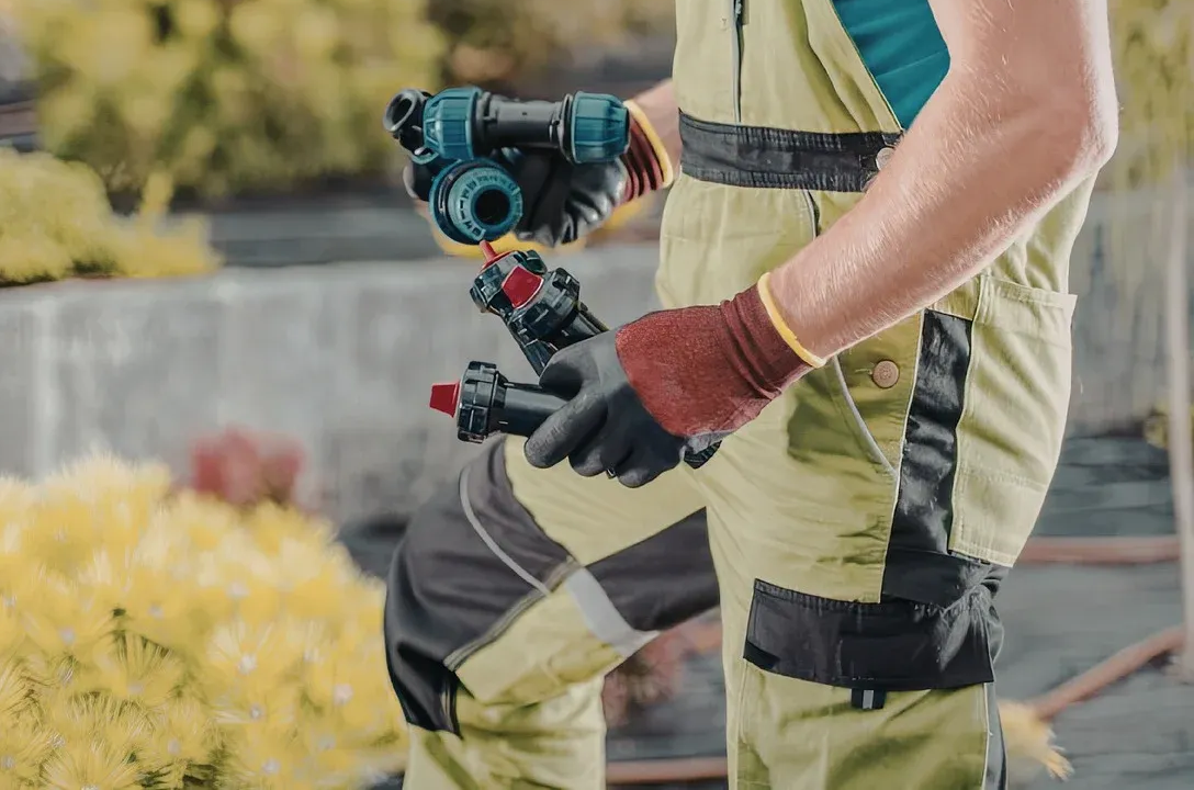 A person in tan work coveralls and gloves holding a specialized irrigation tool in a garden setting.