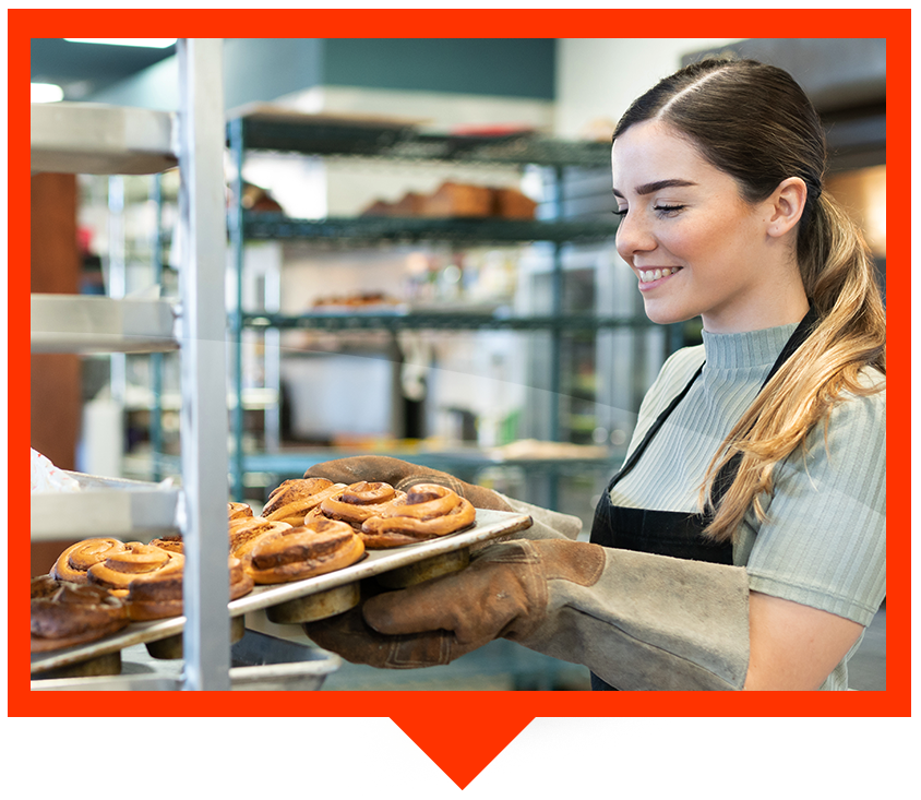 A person in an apron and oven mitts smiles while placing a tray of fresh cinnamon rolls onto a bakery rack.