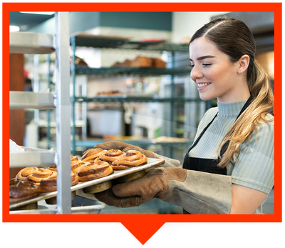 A person in an apron and oven mitts smiles while placing a tray of fresh cinnamon rolls onto a bakery rack.
