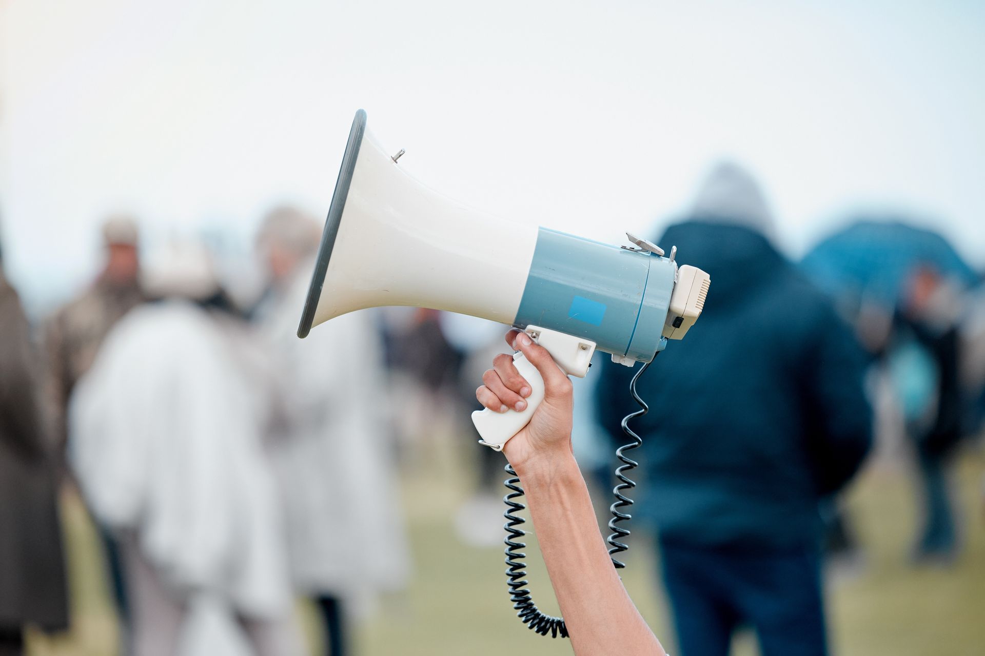 A hand holds a megaphone up toward an out-of-focus crowd outdoors.