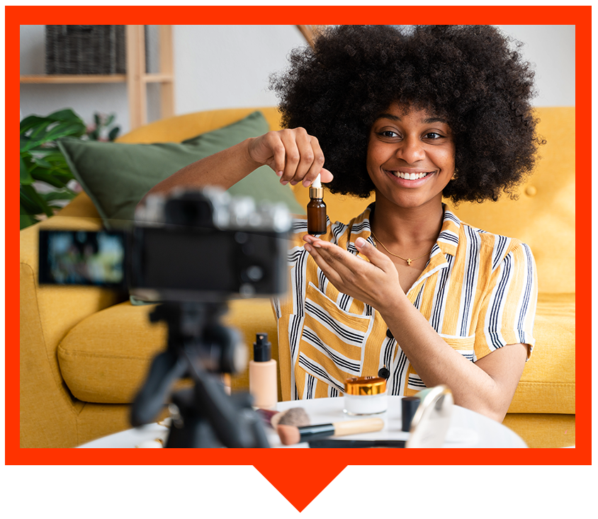 A smiling person recording a beauty product review on camera, holding a small dropper bottle in a bright living room.