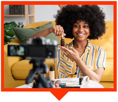 A smiling person recording a beauty product review on camera, holding a small dropper bottle in a bright living room.