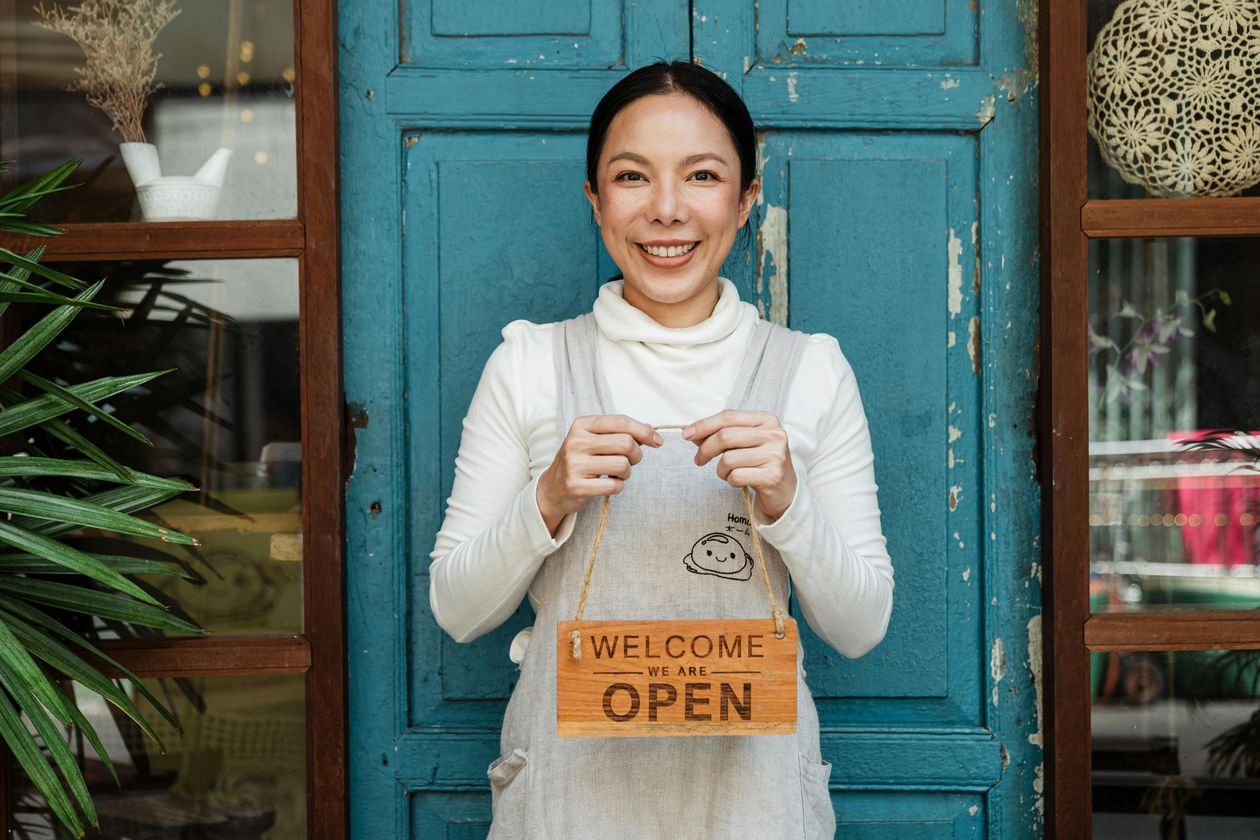 A smiling person stands before a blue door wearing a white apron and holding a wooden 