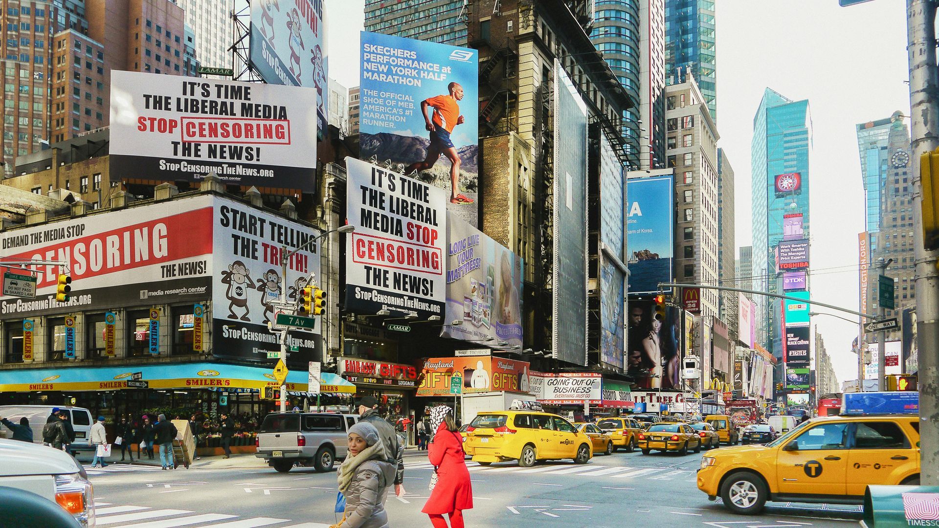 A busy Times Square street scene with yellow taxis and large billboards featuring political slogans.
