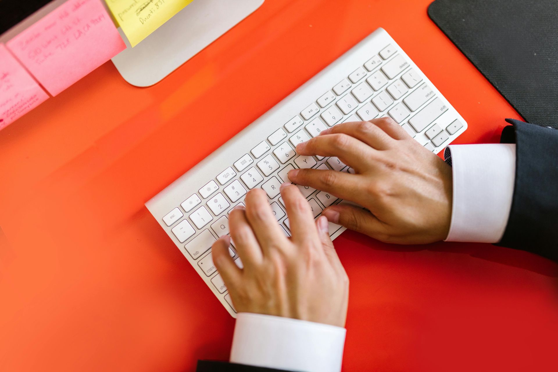 Hands typing on a white keyboard over a bright red desk with sticky notes.