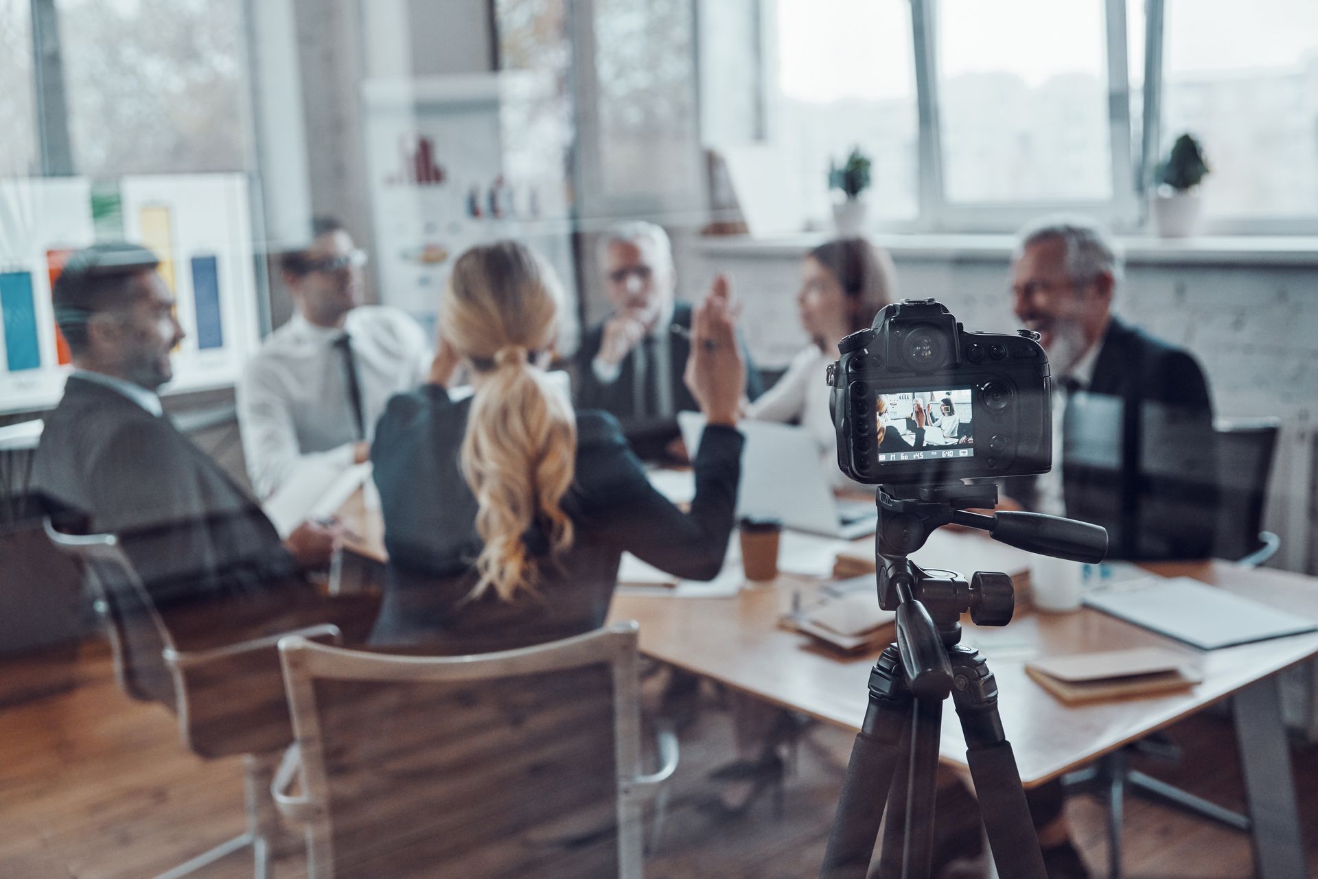 A camera on a tripod faces a group of people sitting around a conference table during a meeting in a bright office.