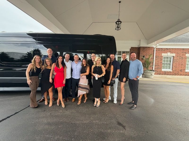 Group of people posing under a covered entrance in front of a black vehicle, with a brick building behind them.
