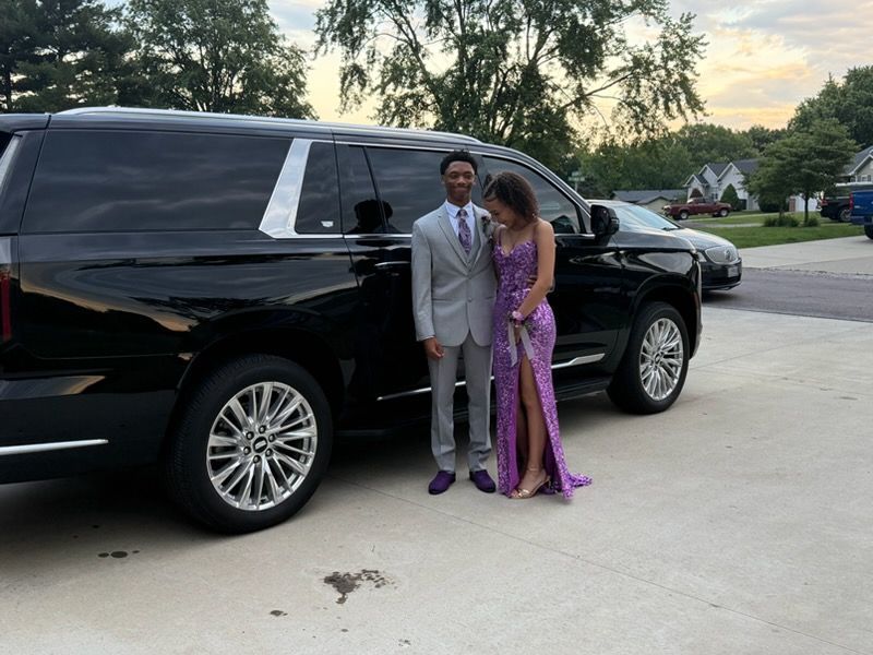 Two people in formalwear standing beside a black SUV on a driveway.