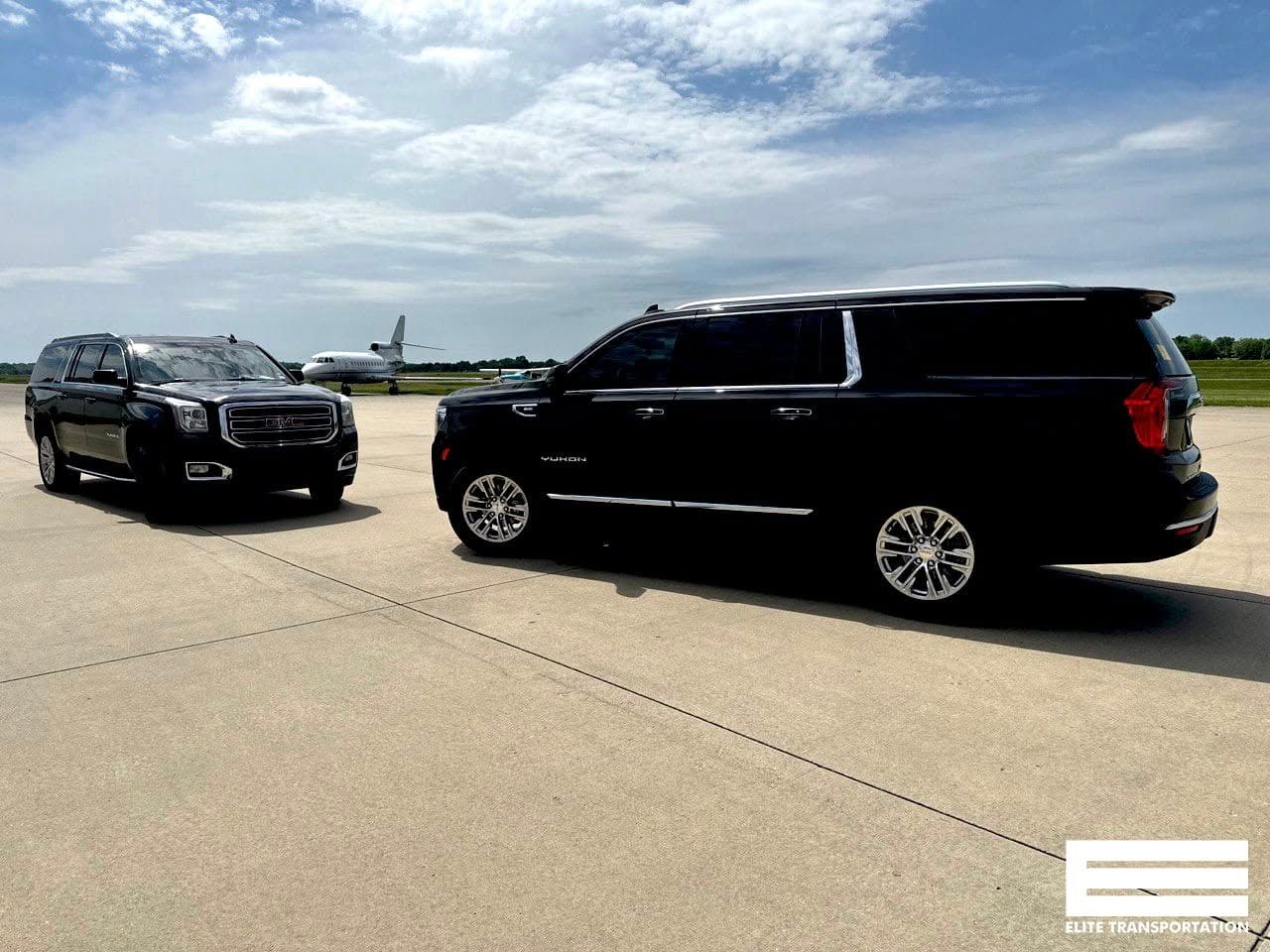 Two black SUVs on a tarmac, plane in the background under a blue sky.
