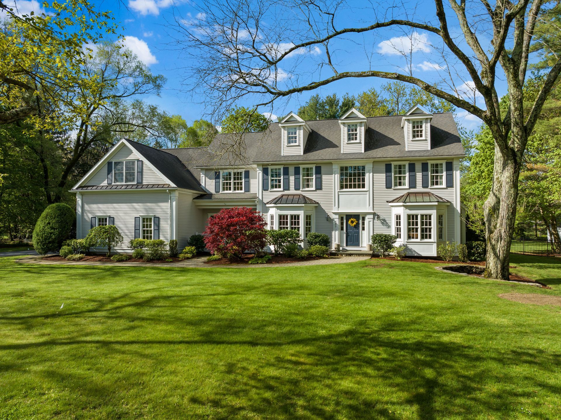 Large white two-story house with dormers, a green lawn, and trees under a blue sky