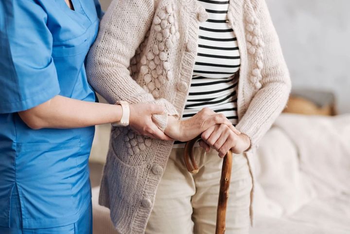 Caregiver holding an elderly person’s hand beside a wooden cane in a bright living room