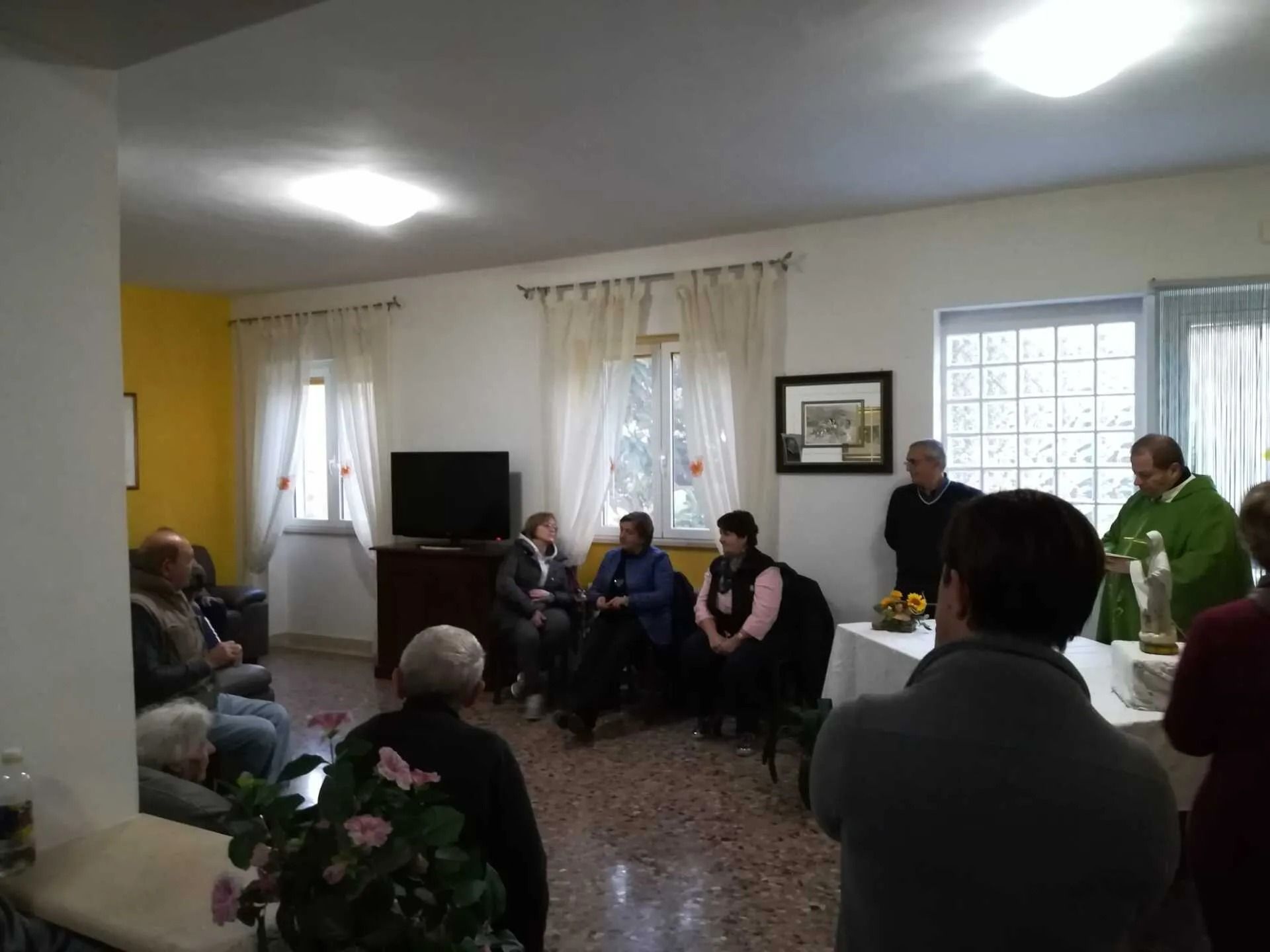 Small group seated in a bright room during a meeting or gathering, with a TV and curtains in the background