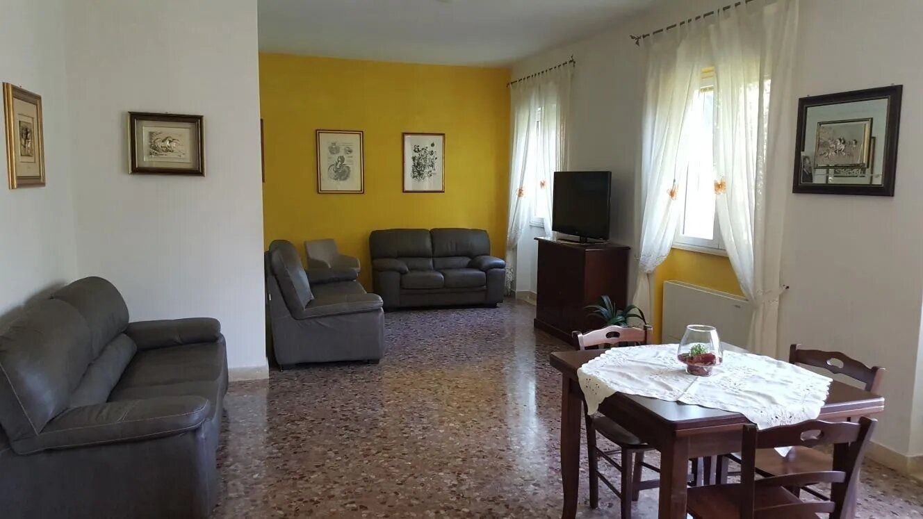 Bright living room with yellow accent wall, gray sofas, large windows, and a dining table by the window