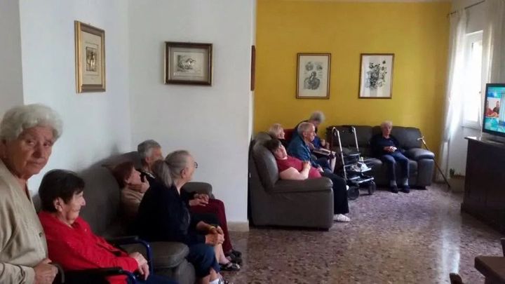 People seated in a living room watching TV, with yellow walls and framed pictures.