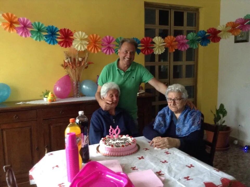 Three people around a birthday cake at a decorated table, with flowers and balloons in a cozy room