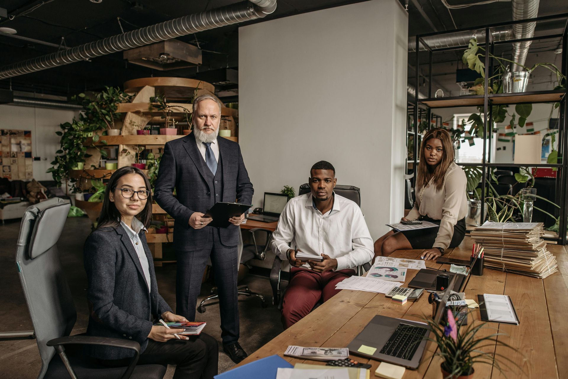 Four coworkers in a modern office, gathered around a wooden table with laptops and documents.