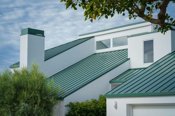 White house with green metal roofs and chimney, partly obscured by trees and shrubs