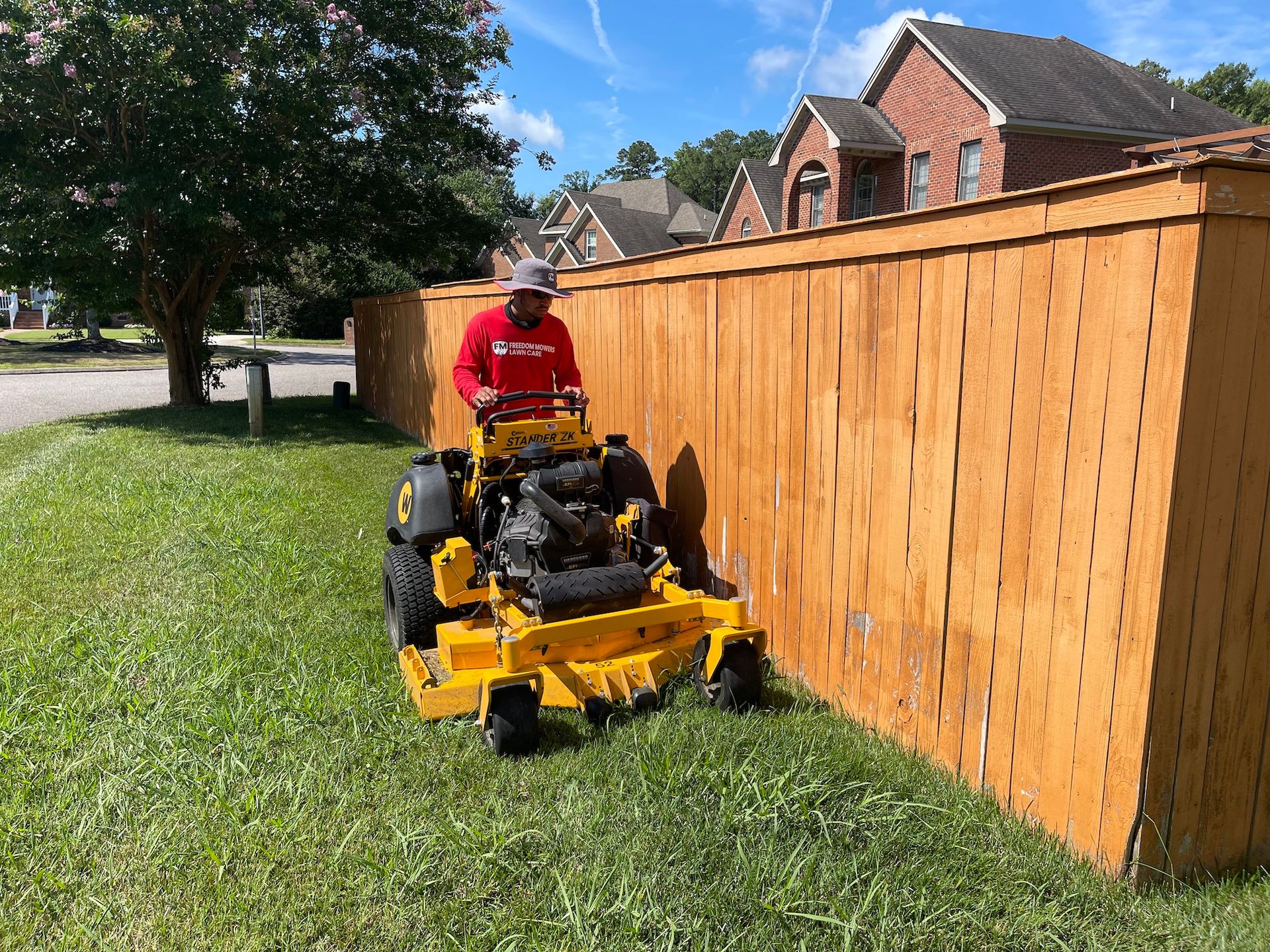 A man is riding a yellow lawn mower next to a wooden fence.